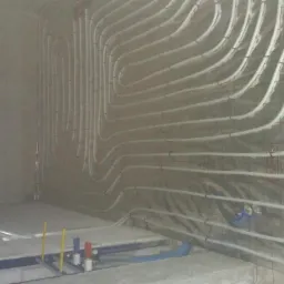 Wewnętrzne ściany i podłoga w trakcie montażu ogrzewania podłogowego i ściennego z widocznymi rurami mocowanymi do izolacji termicznej z metalizowaną folią. Częściowo ułożona podłoga z szarych płyt...