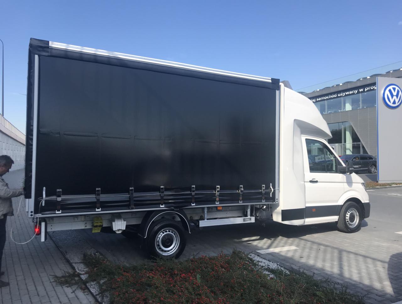 Biały samochód dostawczy z czarną plandeką, stojący przed salonem samochodowym Volkswagen. Widoczny mężczyzna przy tylnej części pojazdu.