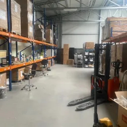 Wnętrze magazynu z regałami wypełnionymi kartonami, wózek widłowy i stanowiska pracy z komputerami w tle.