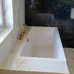 Nowoczesna łazienka z białą wanną obudowaną marmurowymi płytkami z delikatnym złotym wykończeniem krawędzi, bateria wannowa w kolorze złota, ściany wyłożone płytkami imitującymi biały i czarny marmur.