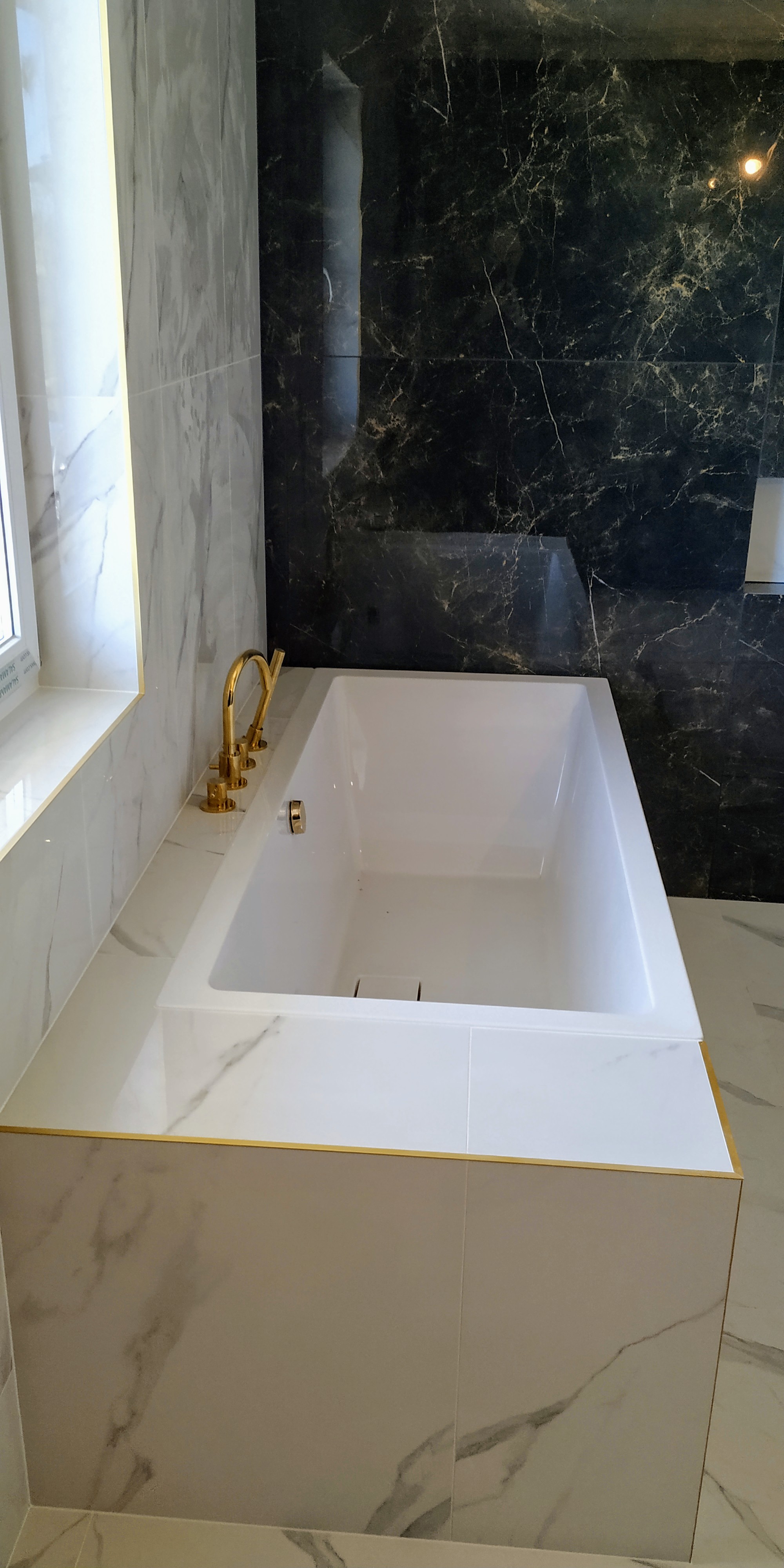 Nowoczesna łazienka z białą wanną obudowaną marmurowymi płytkami z delikatnym złotym wykończeniem krawędzi, bateria wannowa w kolorze złota, ściany wyłożone płytkami imitującymi biały i czarny marmur.