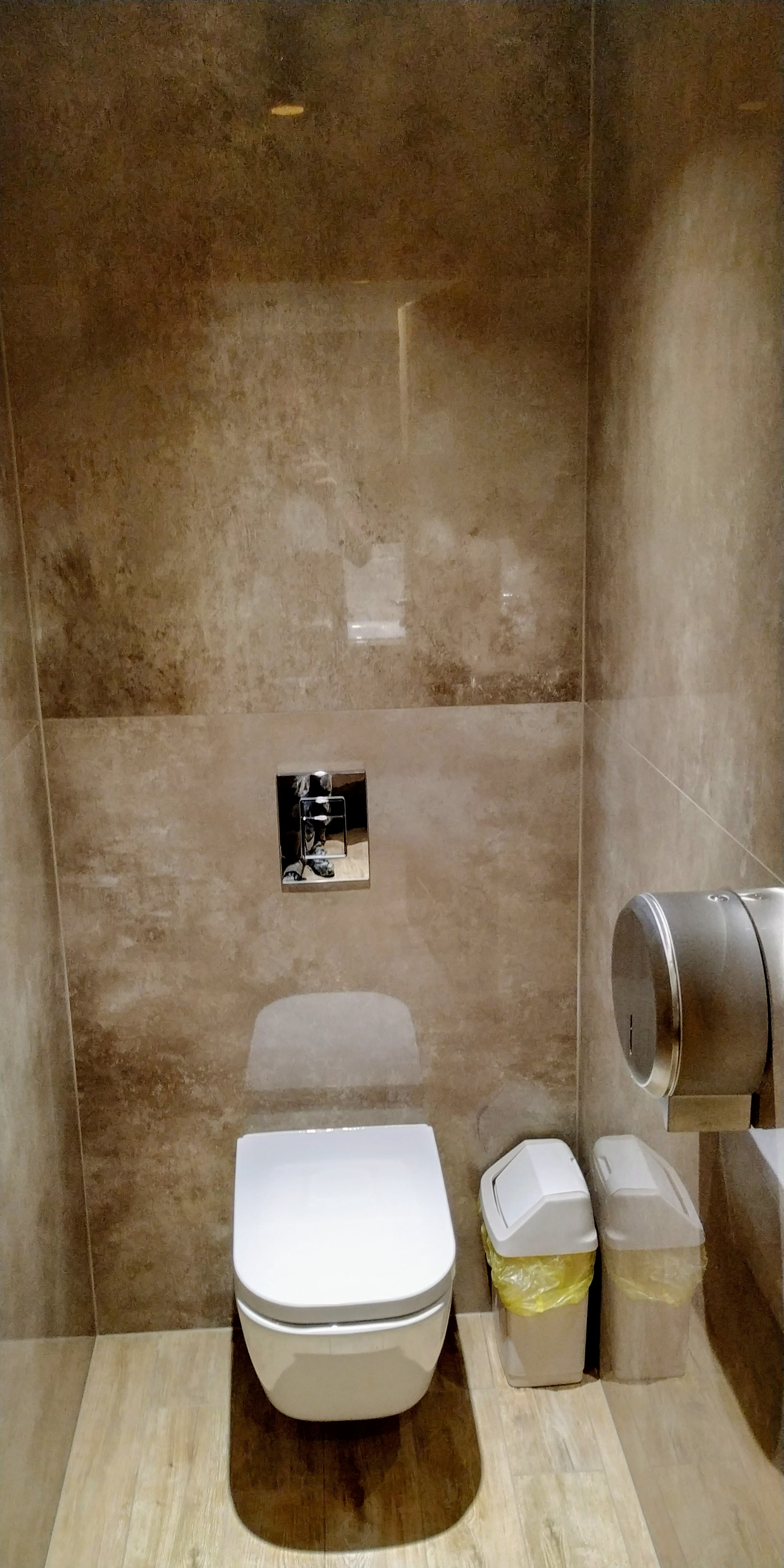 Nowoczesna toaleta podwieszana z beżowymi płytkami imitującymi kamień na ścianach, drewnopodobną podłogą i dwoma koszami na śmieci z żółtymi workami.