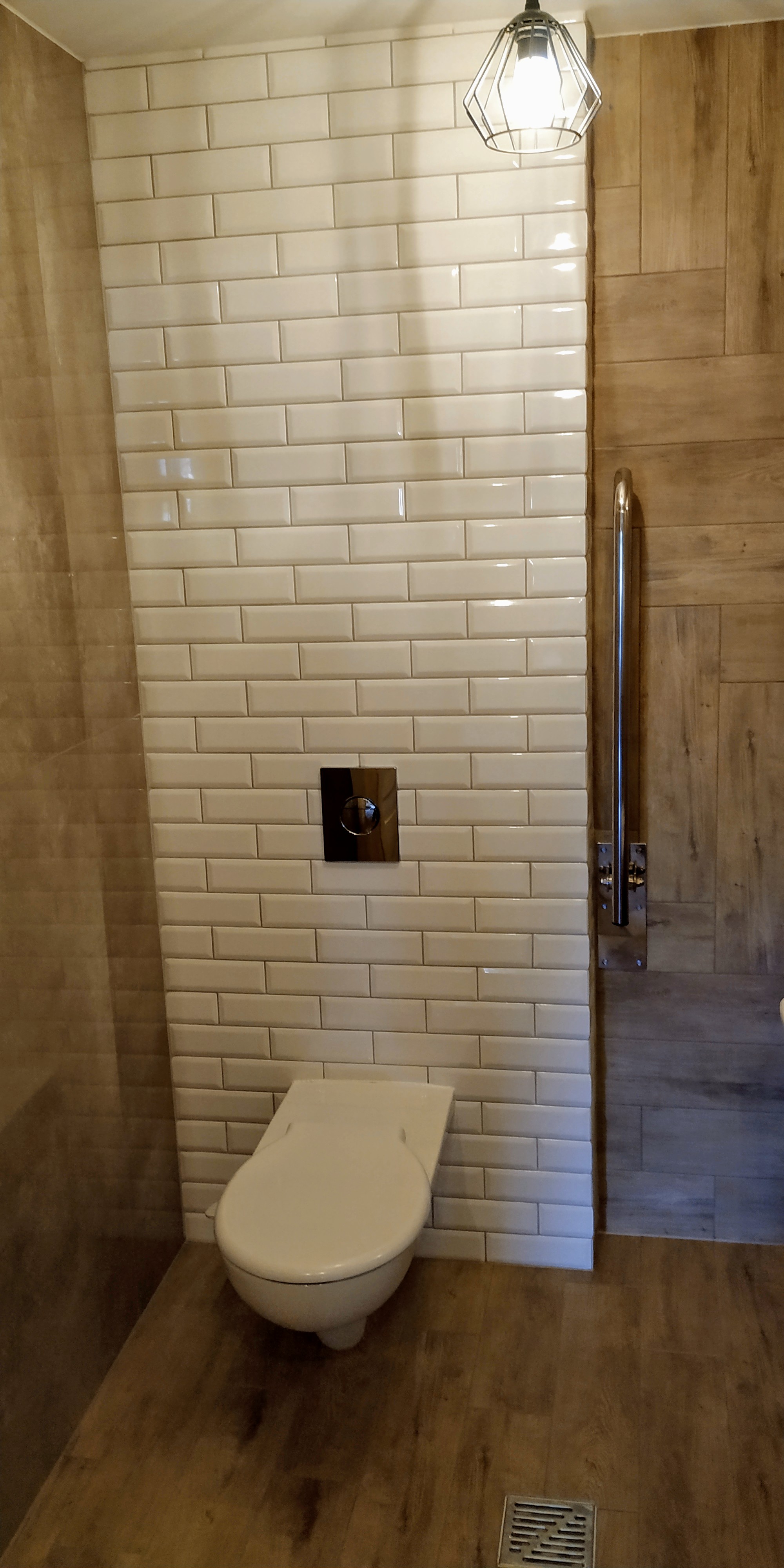 Nowoczesna toaleta z białymi, prostokątnymi płytkami na ścianie, drewnopodobnymi panelami na sąsiedniej ścianie i podłodze, chromowanym uchwytem dla niepełnosprawnych oraz designerską lampą sufitową.