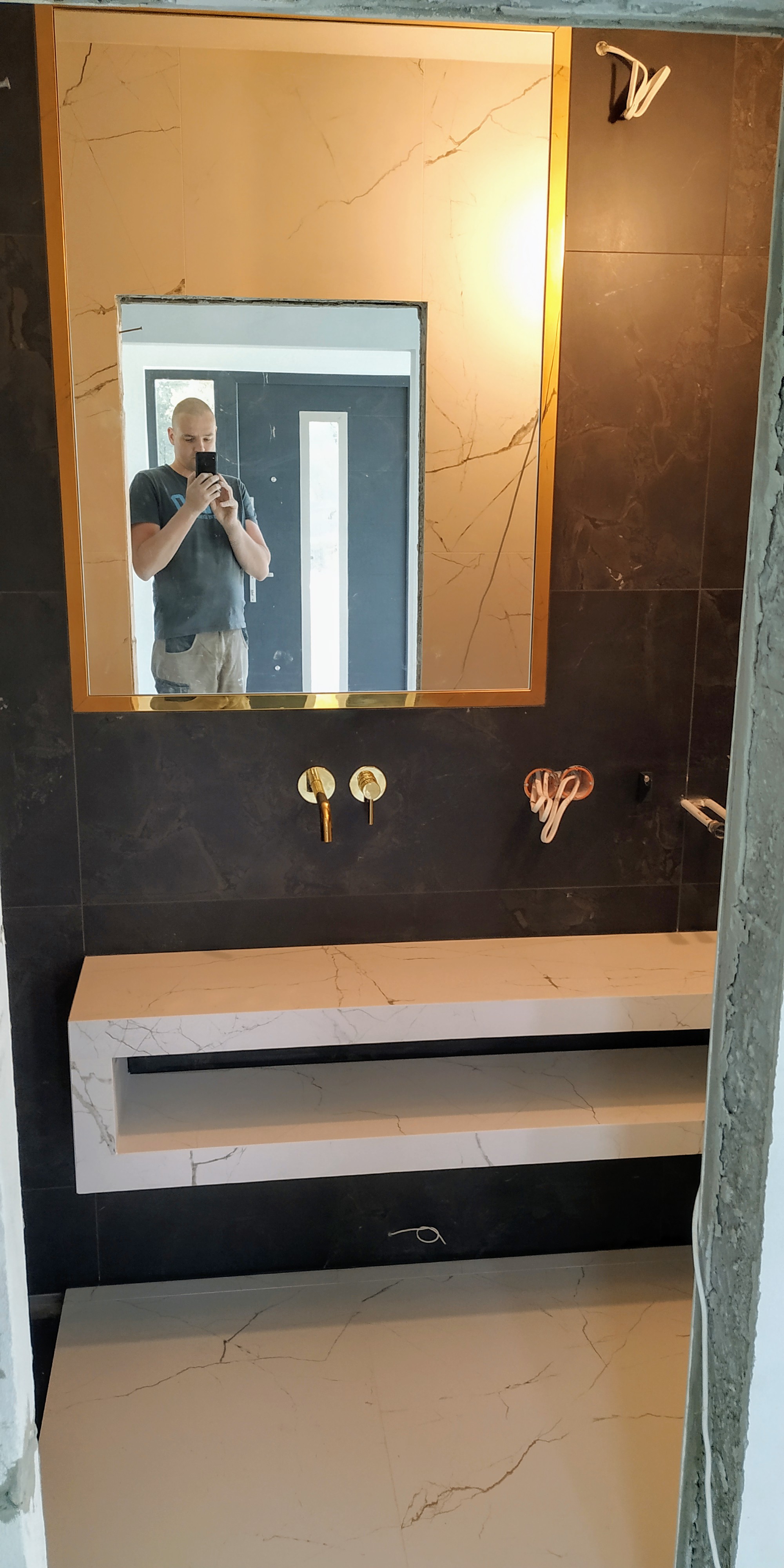 Łazienka w trakcie remontu: wykończone ściany czarnymi i beżowymi płytkami imitującymi marmur, złota armatura, biała marmurowa umywalka z półką, odbicie w lustrze osoby robiącej zdjęcie.