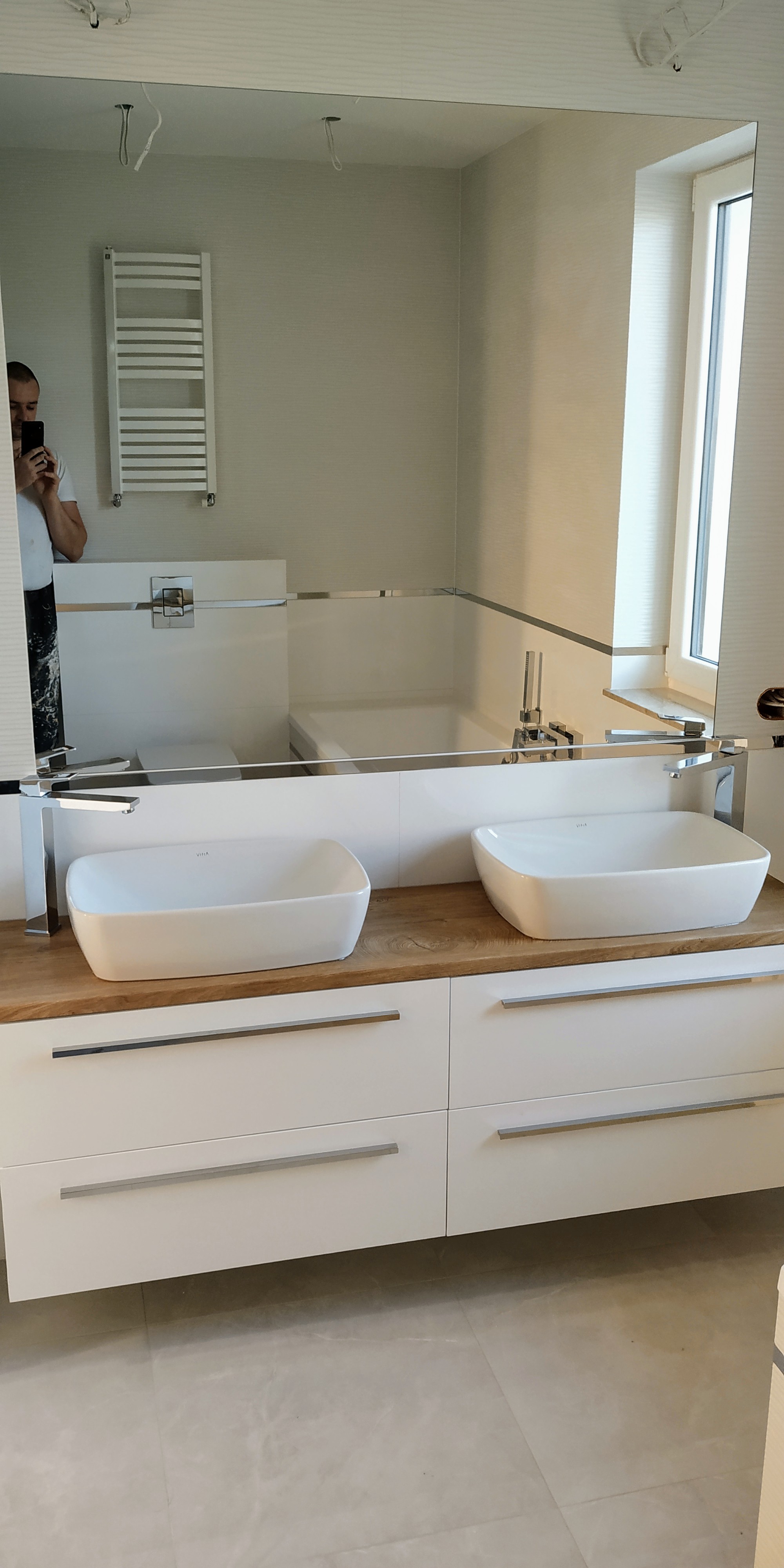 Łazienka z dwiema białymi, prostokątnymi umywalkami nablatowymi na drewnianym blacie i białymi szafkami z szufladami, widoczna w lustrze odbijającym fragment wanny i grzejnika drabinkowego, z osobą...