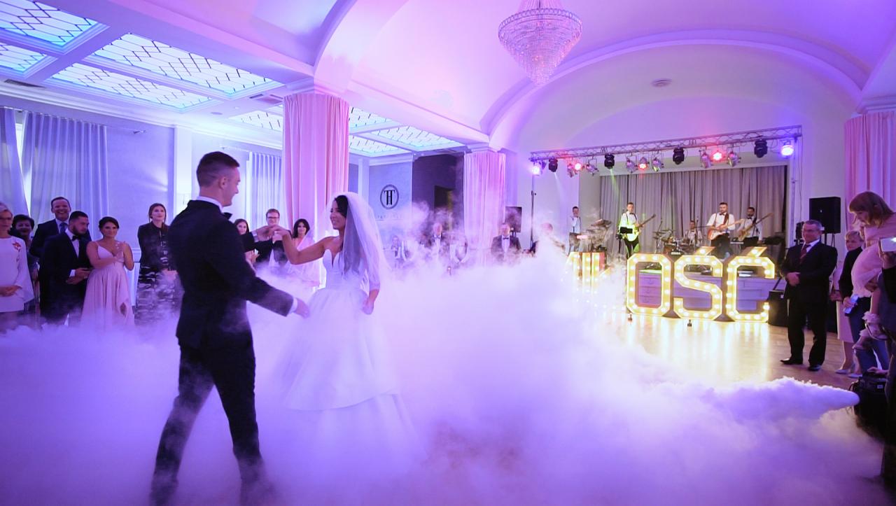 Para młoda w tańcu podczas wesela, otoczona sztucznym dymem, w tle zespół muzyczny i goście.
