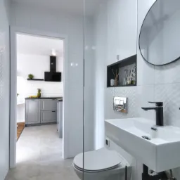 Nowoczesna łazienka z białymi, teksturowanymi płytkami, okrągłym lustrem z czarną ramą, białą umywalką i czarną armaturą, widok na kuchnię w tle przez otwarte drzwi.