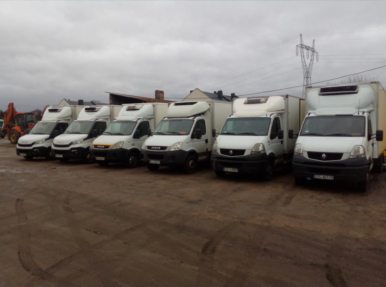 Flota kilku białych samochodów dostawczych chłodni marek Iveco i Renault, zaparkowanych na placu z ubitym piaskiem, w tle słup wysokiego napięcia i budynki.