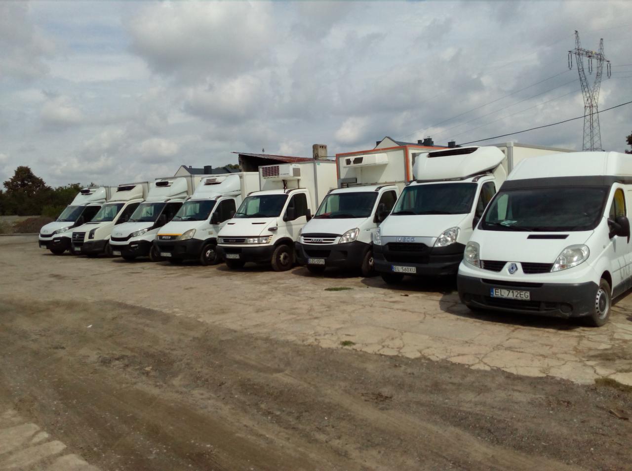 Flota białych samochodów dostawczych z zabudową chłodniczą zaparkowana na placu, widoczne numery rejestracyjne pojazdów.