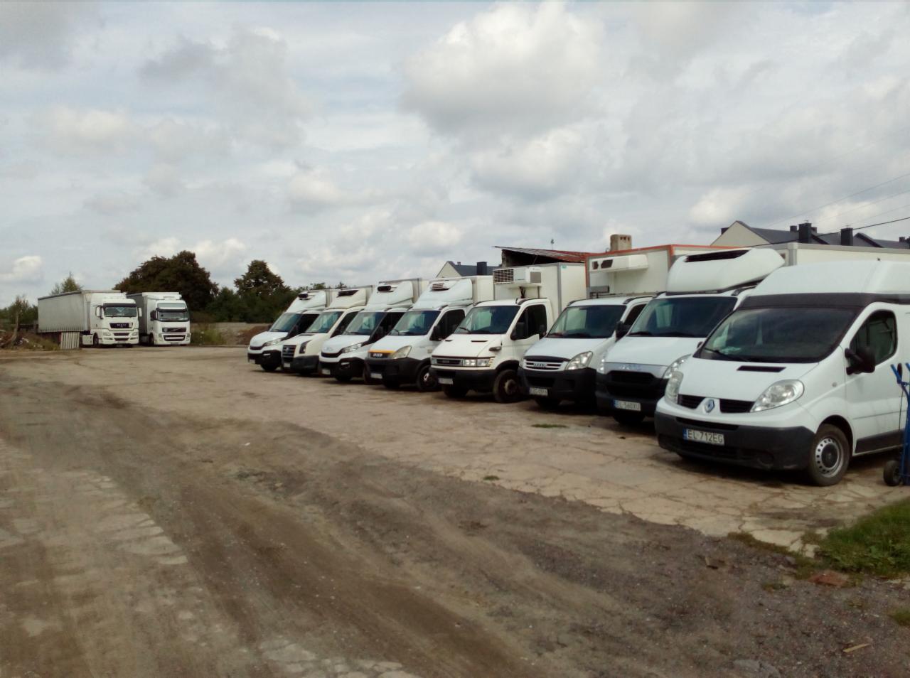 Flota białych samochodów dostawczych z chłodniami i ciężarówek firmy transportowej z Grotnik zaparkowana na placu, widok z boku.