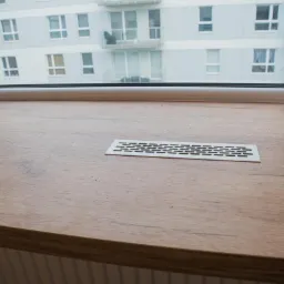 Drewniany blat parapetu z kratkami wentylacyjnymi, widok na blok mieszkalny przez okno.