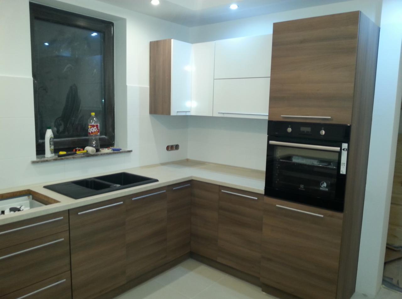 Nowoczesna kuchnia z drewnopodobnymi frontami szafek dolnych i białymi górnymi, czarny zlew dwukomorowy wpuszczony w jasny blat, piekarnik w zabudowie, butelka napoju na parapecie okna.