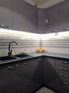 Szara, nowoczesna zabudowa kuchenna z czarnym zlewem i baterią, podświetlana listwą LED nad blatem z szarymi płytkami w poziome pasy. Na blacie widoczny dozownik do mydła i miska z owocami.