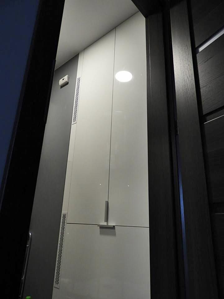 Wysoka, biała szafa wnękowa z metalowymi uchwytami, widoczna z perspektywy wejścia do pomieszczenia, z elementami wentylacyjnymi po bokach.