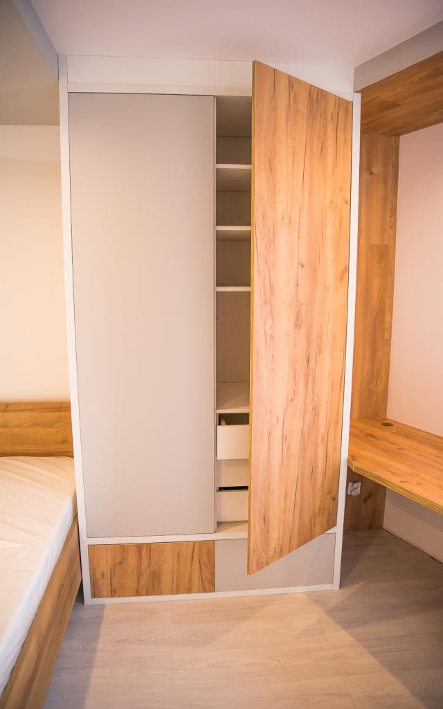 Nowoczesna szafa z przesuwnymi drzwiami, wykończona okleiną drewnopodobną, z otwartym segmentem półek i szuflad, dopasowana do aranżacji sypialni z drewnianym łóżkiem i biurkiem.