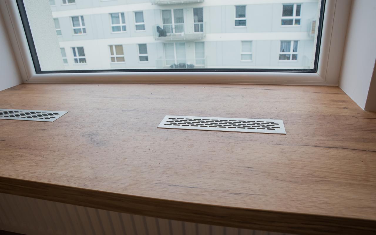 Drewniany blat parapetu z kratkami wentylacyjnymi, widok na blok mieszkalny przez okno.
