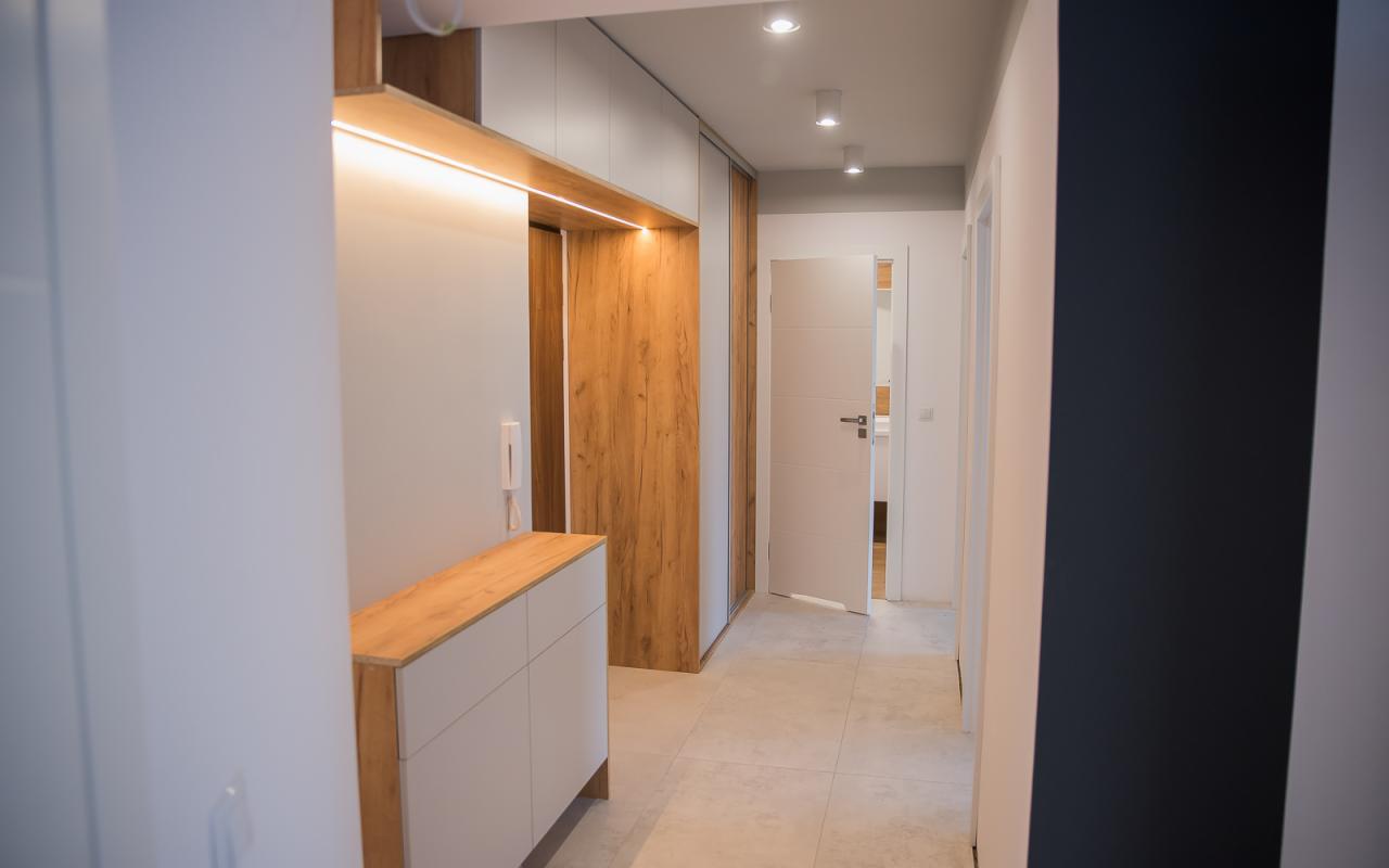 Wnętrze przedpokoju z zabudową meblową: białe szafki z drewnianym blatem, szafa przesuwna z frontem imitującym drewno, oświetlenie LED nad szafkami, jasna podłoga z płytek.