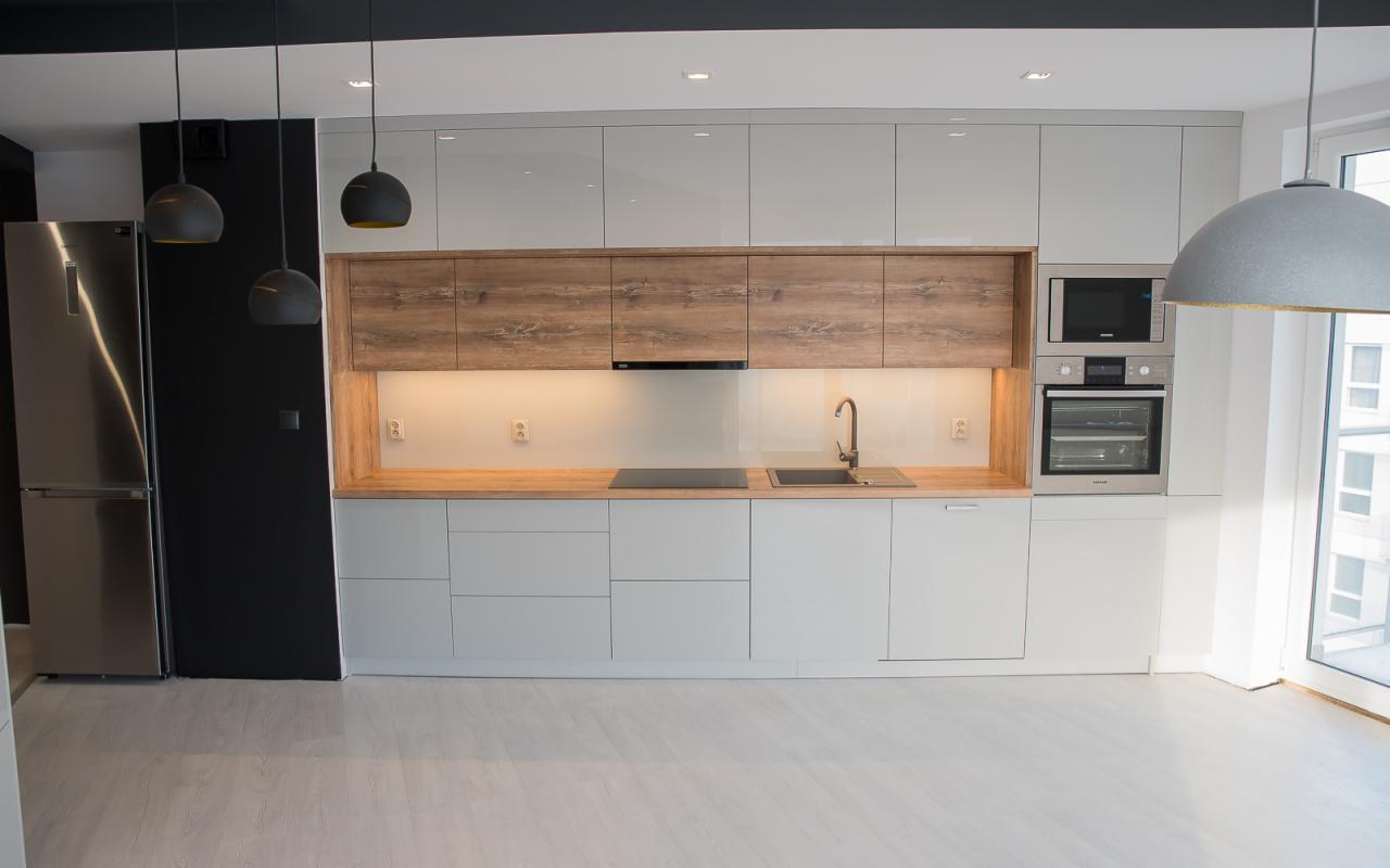 Nowoczesna kuchnia z białymi, lakierowanymi frontami, drewnianymi szafkami górnymi i blatem, oświetlona trzema wiszącymi lampami z czarnymi kloszami.