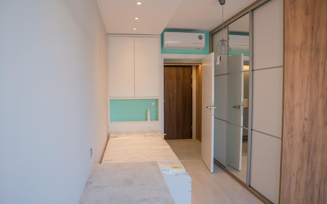 Wnętrze pokoju z zabudową meblową: białe szafki nad łóżkiem z turkusowym panelem, przesuwne drzwi szafy z lustrami oraz drewnopodobnym wykończeniem, klimatyzator na ścianie.