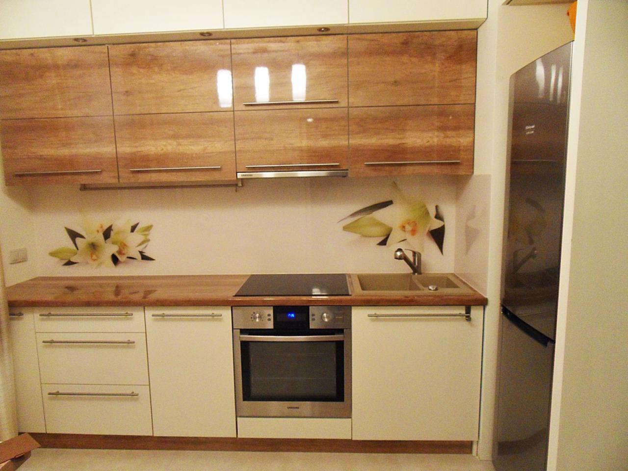 Nowoczesna kuchnia z drewnianymi górnymi szafkami na wysoki połysk, białymi dolnymi szafkami, drewnianym blatem i kwiatowym motywem na ścianie nad blatem, widoczny piekarnik i lodówka.