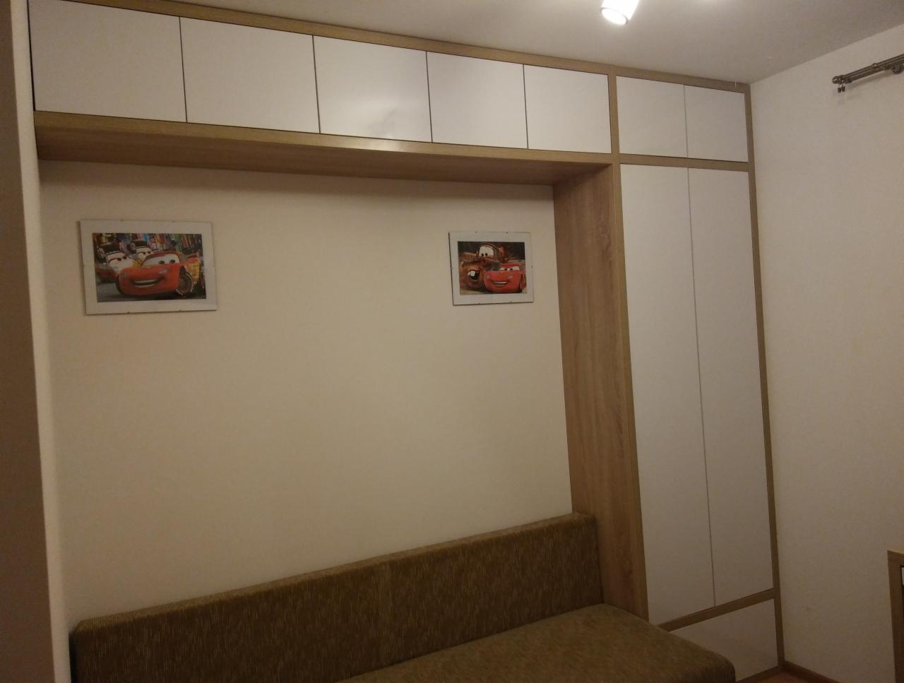 Biało-drewniana szafa wnękowa z górnymi szafkami, pod nią rozkładana sofa i dwa obrazki z motywem bajki 'Auta' na ścianie.