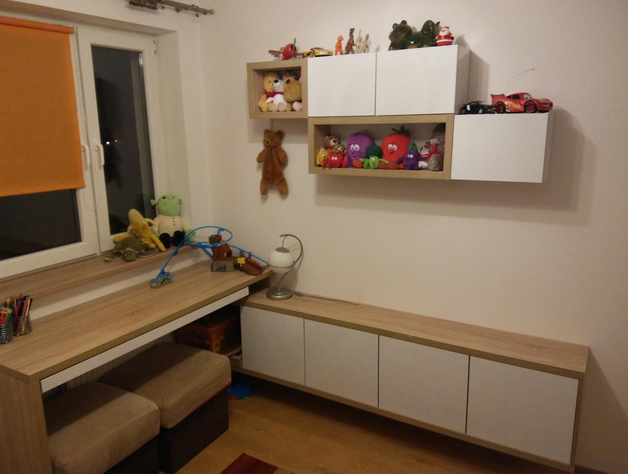 Pokój dziecięcy z meblami: biurko z blatem drewnopodobnym przy oknie z pomarańczową roletą, nad nim wiszące białe szafki z zabawkami, pod biurkiem pufy, a obok długa szafka pod telewizor z białymi...
