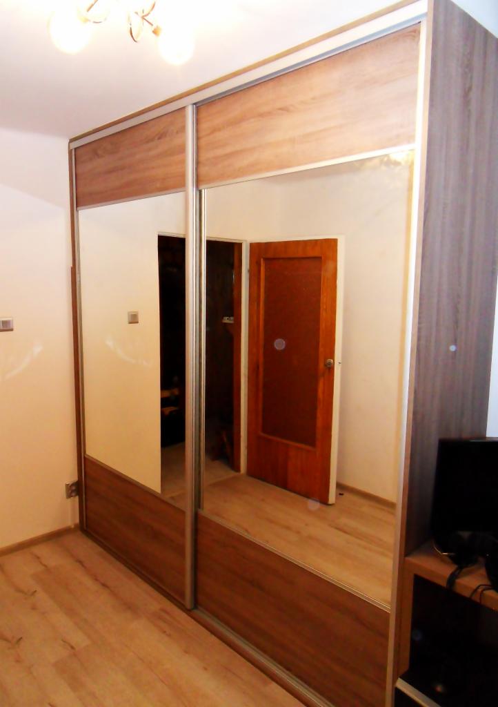 Szafa wnękowa z przesuwanymi drzwiami lustrzanymi, wykończona drewnopodobną okleiną w odcieniach brązu i beżu, z widocznym fragmentem podłogi i drzwi w tle.