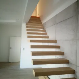 Nowoczesne schody z drewnianymi stopniami bez balustrady, zamontowane na białej ścianie z betonową okładziną, widoczne z dołu, z otwartą przestrzenią między stopniami.