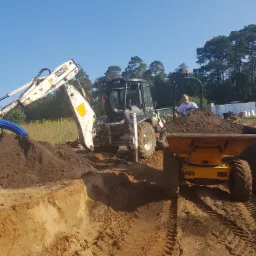 Praca ziemna w toku: koparka z niebieskimi rurami i wywrotka pełna ziemi na tle wykopu, z operatorem w tle. Widoczne drzewa i błękitne niebo.