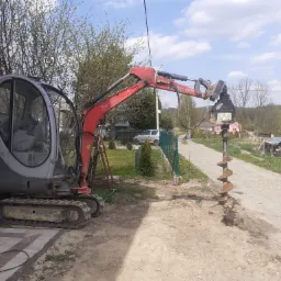Mała koparka z wiertłem świdrowym zagłębiającym się w ziemię, widoczny wykopany otwór, w tle dom jednorodzinny i drzewa.