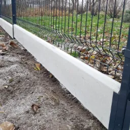 Szary panelowy system ogrodzeniowy z podmurówką betonową, widoczne mocowania słupka, jesienne liście na ziemi.