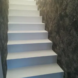 Białe schody w klatce schodowej, ściany wykończone szarą, surową fakturą, widok z dołu.