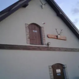 Elewacja budynku z elementami dekoracyjnymi: poroża jelenia, drewniane okiennice z ceglaną obudową, imitacja drewnianej belki, szara ściana i ciemny dach.