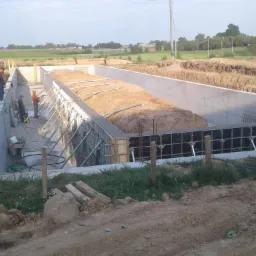 Budowa fundamentów: betonowe ściany szalunkowe zbrojone stalą, wypełnione ziemią, robotnicy przy pracy na placu budowy.