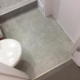 HQRZieliński - Szare płytki podłogowe w łazience, widoczny fragment umywalki i próg kabiny prysznicowej, minimalistyczny styl.