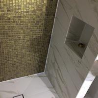 Wykończona łazienka: ściana z mozaiki w odcieniach złota zestawiona z białymi płytami imitującymi marmur, w ścianie wnęka z oświetleniem.