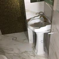 Biała, ceramiczna umywalka na postumencie, z ozdobną, złotą armaturą, na tle ściany wyłożonej mozaiką w odcieniach złota i marmurowej posadzki z odpływem liniowym.