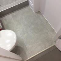 Szare płytki podłogowe w łazience, widoczny fragment umywalki i próg kabiny prysznicowej, minimalistyczny styl.