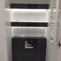 Nowoczesna łazienka z czarnymi płytkami na zabudowie WC i półkach, podświetlane LED, białe ściany i grzejnik drabinkowy.