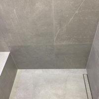 Szara, matowa podłoga i ściany wyłożone wielkoformatowymi płytkami ceramicznymi imitującymi marmur z delikatnym użyleniem. Widoczny odpływ liniowy w podłodze i fragment białej umywalki.