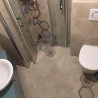 Beżowa łazienka z heksagonalnymi płytkami na podłodze, kabina prysznicowa ze srebrnymi profilami, biała toaleta i umywalka, dekoracyjne płytki w kształcie plastra miodu na ścianie.