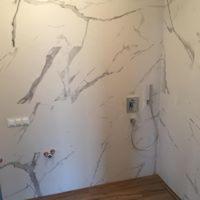 Ściana łazienki wyłożona białymi płytami imitującymi marmur z szarymi żyłami, widoczna armatura prysznicowa i gniazdka elektryczne, podłoga z drewnopodobnych paneli.