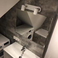 Nowoczesna łazienka z szarą, betonową ścianą, umywalką z chromowaną baterią i dużym lustrem odbijającym pralkę oraz fragment wanny.