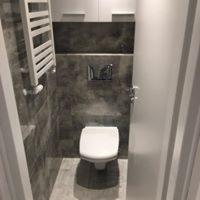 Nowoczesna łazienka z szarą, marmurkową okładziną ścienną, podwieszana toaleta, biały grzejnik drabinkowy i zabudowa szafek nad stelażem WC.