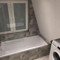 Wykończona łazienka na poddaszu: biała wanna zabudowana szarą, imitującą kamień glazurą, białe okno w ramie z widocznym fragmentem pralki. Skośny sufit.