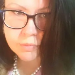 Portret kobiety w okularach z perłowym naszyjnikiem, skierowanej w stronę obiektywu, z widocznym cieniem na twarzy.