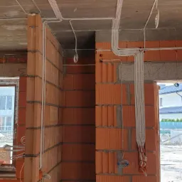 Instalacja elektryczna w surowym budynku z czerwonej cegły, widoczne kable prowadzone po suficie i ścianach, okno z widokiem na sąsiedni dom.