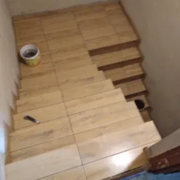 Schody wyłożone płytkami imitującymi drewno, widoczny kubeł z zaprawą i nóż do tapet