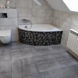 Nowoczesna łazienka na poddaszu z szarymi płytkami, mozaiką na obudowie wanny narożnej, wiszącą toaletą i białym grzejnikiem żeberkowym pod oknem dachowym.