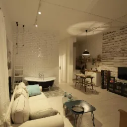 Przestronne wnętrze mieszkania w stylu loft z wanną wolnostojącą, ceglaną ścianą i drewnianymi panelami, oświetlone reflektorami punktowymi i lampą wiszącą.